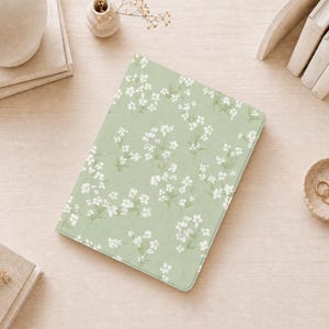 May include: A rectangular notebook with a sage green cover featuring a repeating pattern of small white flowers and thin green stems. The notebook is positioned at an angle, displaying a flat, smooth surface with a consistent floral print design.