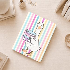 Puede incluir: Un cuaderno rectangular con una oca blanca que lleva gafas redondas y sostiene una pila de tres libros. El fondo consiste en rayas verticales en tonos pastel de rosa, amarillo y azul. La cubierta tiene un acabado mate y suave.