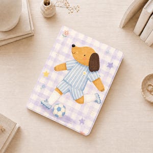 Könnte beinhalten: Ein rechteckiges Notizbuch mit einem hellvioletten und weißen Vichy-Karo-Hintergrund. Das Cover zeigt einen beigen Hund in einem hellblau-weiß gestreiften Fußballtrikot, der auf einen schwarz-weißen Fußball zuläuft. Kleine gelbe Sterne und winzige Blumen sind über das Cover verteilt.