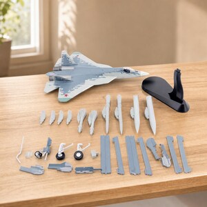 May include: A gray and white model fighter jet with a pixelated camouflage pattern and red star insignias on the wings. The set includes a black display stand, several detachable white missiles, landing gear components, and various gray plastic parts.