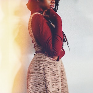 May include: A woman wearing a maroon turtleneck sweater and a brown crocheted dress. The dress has a flared skirt and a belt.