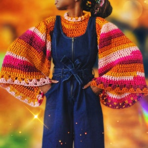 Puede incluir: Una mujer lleva una blusa de crochet a rayas con mangas acampanadas en tonos de rosa, naranja y marrón. La blusa se lleva sobre un mono vaquero con cremallera y cinturón.