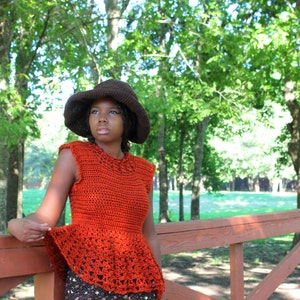 May include: An orange crochet top with a peplum design and a brown hat. The sleeveless top has a detailed neckline and a flared bottom. The hat has a wide brim. The background features trees and a wooden bridge.