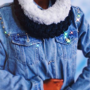 May include: A denim jacket with gold studs on the shoulders and a fluffy, two-tone scarf. The scarf has a white faux fur trim and a dark blue knitted center. The jacket has two front pockets and a button closure.
