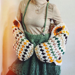 K&ouml;nnte beinhalten: Ein gr&uuml;ner H&auml;kelrock mit einem wei&szlig;en und gelben H&auml;keltop. Das Oberteil hat ein Granny-Square-Muster und wird &uuml;ber einem beigen Rollkragenpullover getragen.