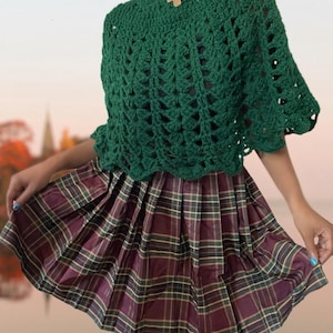 May include: A green crocheted poncho with a scalloped edge and a wooden mushroom button. The poncho is paired with a plaid skirt.