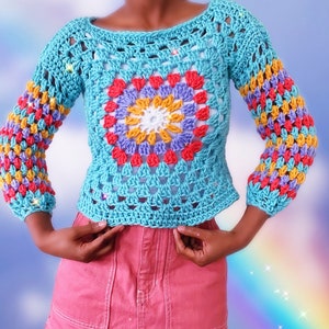 Puede incluir: Un top de crochet con un diseño de granny square colorido en tonos de azul, rosa, amarillo y morado. El top tiene mangas largas y un ajuste holgado.
