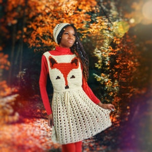 May include: A cream-colored crochet dress with a fox face design on the front. The fox face is made of orange and brown yarn. The dress has a matching cream-colored hat. The person is wearing a red turtleneck and leggings.
