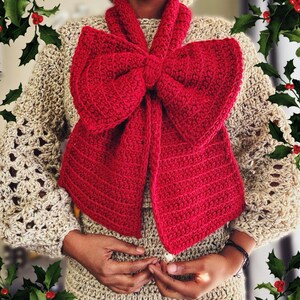 May include: A handmade, red crochet scarf in a bow shape, worn over a beige crochet sweater. The sweater has openwork sleeves. The image is framed with holly and red berries, suggesting a holiday theme.