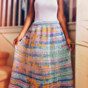 May include: A colorful, handmade crochet skirt in shades of pink, blue, yellow, and orange. The skirt has a wide, flowing design and is paired with a white ribbed tank top. The skirt appears to be knee-length.