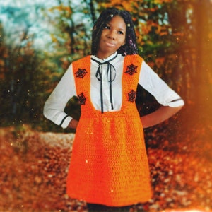 May include: A young person wearing a white shirt with a black bow and an orange crocheted dress with black spiderweb designs. The dress has a pinafore style and is worn over the white shirt.