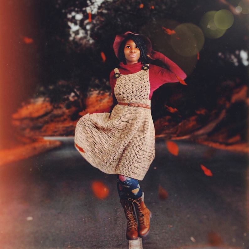 May include: A woman wearing a crocheted dress with a brown belt, a burgundy sweater, and brown boots. She is standing in a forest with fallen leaves on the ground.