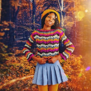 May include: A woman wearing a colorful striped crochet top with a yellow crocheted hat. She is standing in a forest setting with fall foliage.
