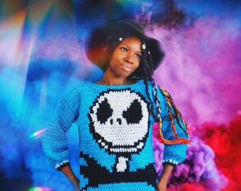 Puede incluir: Una persona joven lleva un suéter de crochet azul con un diseño de Jack Skellington blanco y negro. El suéter tiene mangas largas y un ajuste holgado. La persona lleva un sombrero negro y tiene trenzas largas.