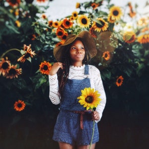 May include: A young person wearing a blue crocheted overall dress with a white lace top and a brown straw hat. They are holding a sunflower in front of a field of sunflowers.