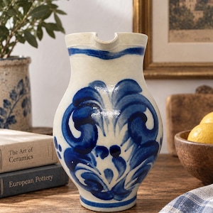 Op de afbeelding: Een keramische kan met een crèmekleurige basis en handgeschilderde kobaltblauwe florale krulpatronen. De kan heeft een brede, ronde buik, een smalle hals en een duidelijke schenktuit, met blauwe horizontale banden rond de rand en de voet.
