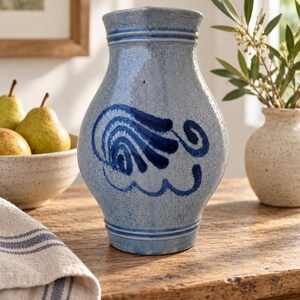 May include: A light blue ceramic pitcher with a speckled finish and a dark blue hand-painted swirl design on the front. The pitcher features a flared rim, a tapered neck, and two horizontal embossed bands near the top and bottom edges.