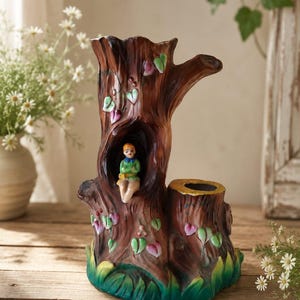 May include: A brown ceramic tree stump planter featuring a hollow opening with a small seated figure inside. The exterior is decorated with painted green and pink leaves and vines, with a green base and a secondary cylindrical opening on the right.