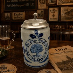 Op de afbeelding: Een grijze keramische bierpul met een metalen deksel, voorzien van een blauw cirkelvormig embleem met een boomillustratie en de tekst Die Alten Deutschen 1889 Bamberg. De pul heeft een cilindrisch lichaam met een gladde afwerking en een knop.