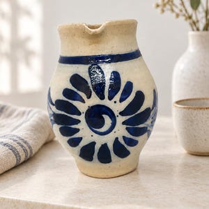 Op de afbeelding: Een crèmekleurige keramische kan met een handgeschilderd donkerblauw bloemmotief en een centraal spiraalontwerp. De kan heeft een brede, ronde vorm, een korte hals en een kleine schenktuit, afgewerkt met een glanzende glazuurlaag.