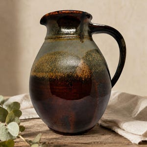 May include: A ceramic pitcher with a rounded body and a curved handle. The glaze features a gradient of deep brown at the base, transitioning into a mottled olive green and grey midsection, with a dark, glossy rim and a small pouring spout.
