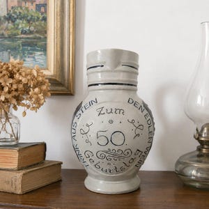 Puede incluir: Una jarra de gres cerámico de color gris claro con un borde azul oscuro y texto decorativo que dice Zum 50. Geburtstag y Trink aus Stein den edlen Wein. El recipiente tiene un cuerpo redondeado, un cuello estrecho y un pequeño pico vertedor.