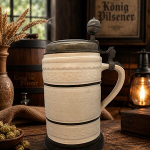 Op de afbeelding: Een crèmekleurige keramische bierpul met een metalen deksel met duimrust, een stevig handvat en horizontale banden met reliëf bloemmotieven. De pul heeft een donkere metalen voet en een cilindervormig lichaam met een matte textuur.