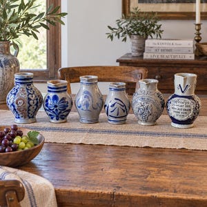 Może przedstawiać: Kolekcja sześciu ceramicznych dzbanków ustawionych w rzędzie. Każde naczynie posiada unikalne szaro-niebieskie szkliwo, a niektóre z nich zdobią wypukłe wzory kwiatowe, motywy winogron oraz niemieckie napisy na ich zaokrąglonych korpusach.