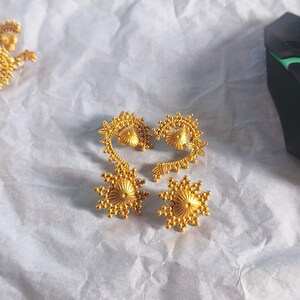 Peut inclure: Un ensemble de bijoux dorés composé de deux boucles d'oreilles rondes assorties avec un motif en rayons de soleil et deux grandes boucles d'oreilles incurvées présentant des motifs filigranés, des bords perlés et un dôme central.