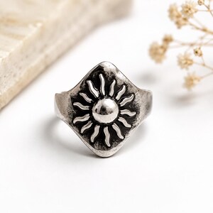 Op de afbeelding: Een zilverkleurige metalen ring met een ruitvormig voorvlak en een verhoogd, gepolijst zonmotief in het midden. De achtergrond van het zonontwerp is donkerder gemaakt met een geoxideerde afwerking, wat zorgt voor contrast met de stralen.