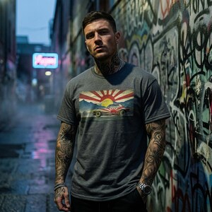 Könnte beinhalten: Ein anthrazitfarbenes Kurzarm-T-Shirt mit einem rechteckigen Grafikdruck. Das Design zeigt ein Oldtimer-Auto vor einer stilisierten aufgehenden Sonne mit roten Strahlen, einer gelben Sonnenscheibe und blauen Bergsilhouetten im Hintergrund.