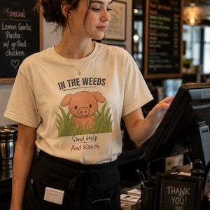 May include: A cream-colored short-sleeve t-shirt featuring a graphic of a pink pig peeking out of green grass. The text above the pig reads IN THE WEEDS and the text below reads Send Help And Ranch in black and red lettering on the front of the shirt.