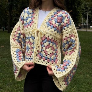 Puede incluir: Un cárdigan de ganchillo de manga larga confeccionado con cuadrados de la abuela en color crema, azul claro y rosa empolvado. La prenda presenta un escote en pico, mangas acampanadas y un cierre frontal de lazo en hilo crema, sobre una camiseta blanca.
