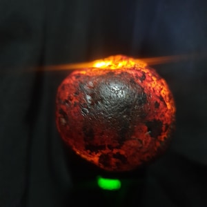 May include: A rounded, textured stone with a dark, mottled surface in shades of deep red and black. A bright, warm orange light glows from behind the top edge of the stone, creating a luminous effect that highlights the uneven, rugged surface.