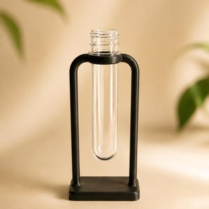 May include: A clear glass test tube suspended within a matte black metal frame. The frame consists of a rectangular base and two vertical arms that curve inward at the top to hold the cylindrical glass vessel securely in an upright position.