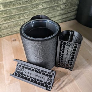 May include: A black cylindrical container with a textured exterior and a clear top rim. Beside it are two black, semi-cylindrical attachments featuring a hexagonal honeycomb pattern and the embossed text Green Haven on each piece.