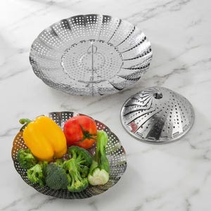 May include: Two stainless steel vegetable steamer baskets with a collapsible petal design. One basket is fully expanded and holds a yellow bell pepper, a red tomato, broccoli florets, and bok choy. The second basket is shown in a folded position.