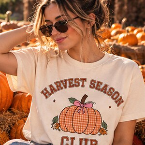 May include: A cream-colored short-sleeve t-shirt featuring the text "HARVEST SEASON CLUB" in brown block letters. The center graphic displays an orange gingham-patterned pumpkin with a pink bow, green leaves, and small orange flowers.
