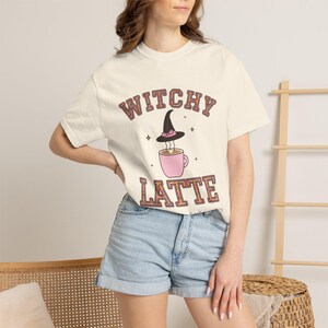 May include: A cream-colored short-sleeve t-shirt featuring the text "WITCHY LATTE" in a brown and orange patterned font. The graphic includes a pink coffee mug with steam rising toward a black witch hat with a pink band and small star accents.