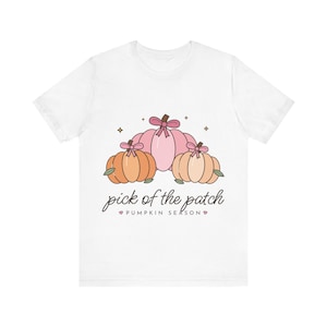May include: A white short-sleeve t-shirt featuring a graphic of three pumpkins in shades of pink and orange, each topped with a small pink bow. Below the pumpkins, the text reads "pick of the patch" and "PUMPKIN SEASON" with small heart icons.