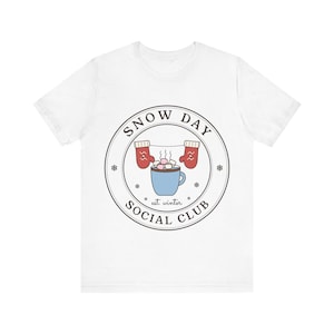 May include: A white short-sleeve t-shirt featuring a circular graphic with the text SNOW DAY SOCIAL CLUB and est. winter. The design includes a light blue mug filled with marshmallows, two red mittens hanging on a string, and small snowflakes.