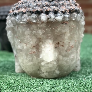 Può includere: Una scultura a testa di Buddha traslucida color bianco sporco, realizzata con frammenti di pietra chiari e bianchi incastonati. La parte superiore della testa presenta noduli scuri e arrotondati accentuati da scaglie metalliche color rame.