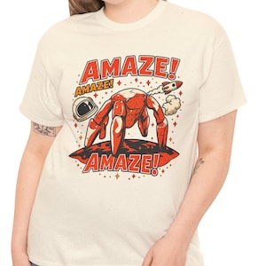 May include: A cream-colored short-sleeve t-shirt featuring a graphic of a red crab in a space suit with a rocket, an astronaut helmet, and the text "AMAZE!" printed in red above and below the illustration on a dark red planet surface.