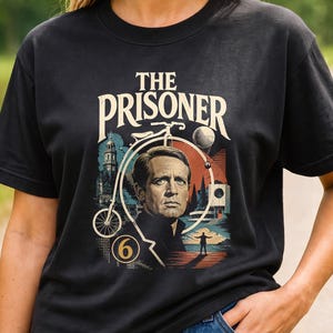 Peut inclure: Un t-shirt noir à manches courtes avec une impression graphique portant le texte THE PRISONER. Le motif comprend le portrait d'un homme, un grand-bi, une sphère, un bâtiment et le chiffre 6, le tout sur un fond aux tons colorés.