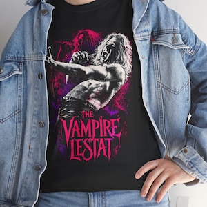 May include: A black t-shirt featuring a graphic print of a long-haired figure singing into a microphone, with the text "THE VAMPIRE LESTAT" in bold pink lettering. The design includes vibrant pink and purple splatters and dark silhouettes.