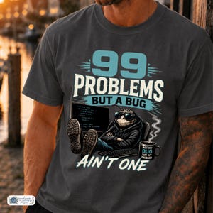 Könnte beinhalten: Ein anthrazitfarbenes T-Shirt mit einer Grafik eines Insekts mit Sonnenbrille und Kapuzenpullover, das vor einem Computer sitzt. Der Text lautet 99 PROBLEMS BUT A BUG AIN'T ONE, neben der Tastatur steht eine Tasse mit der Aufschrift NOT MY BUG NOT MY PROBLEM.