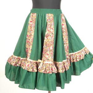May include: A forest green skirt featuring vertical panels of a multi-colored fruit and floral print. The skirt has an elastic waistband, a ruffled hemline trimmed with a thin gold ribbon, and a small decorative bow detail on the front panel.