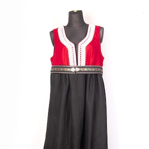 Puede incluir: Vestido tradicional con corpiño de terciopelo rojo y falda negra. El corpiño presenta un ribete de encaje blanco a lo largo del escote y una banda bordada decorativa en la cintura. El vestido se exhibe en un maniquí negro.