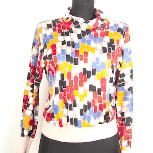 Op de afbeelding: Een top met lange mouwen, een hoge kraag en een elastische zoom. De stof heeft een herhalend patroon van kleine, rechthoekige penseelstreekvormen in levendige tinten rood, geel, blauw, zwart en wit tegen een lichte achtergrond.