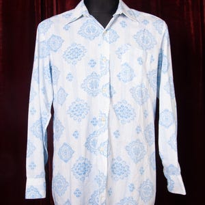 Puede incluir: Una camisa blanca con un estampado floral azul claro. La camisa de manga larga tiene un cuello clásico y un bolsillo en el pecho. El patrón es un diseño repetido de flores estilizadas y formas geométricas.